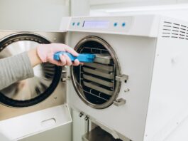A close-up of a hand placing a tray on a rack in a freeze-drying machine. The person uses blue tongs to touch the tray.