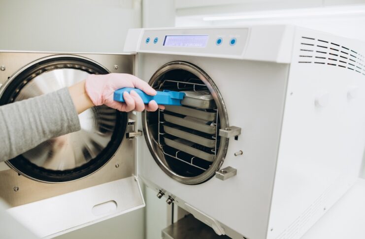 A close-up of a hand placing a tray on a rack in a freeze-drying machine. The person uses blue tongs to touch the tray.