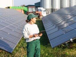 A female farmer wearing a green hat and pants stands between two rows of solar panels in a farm field.