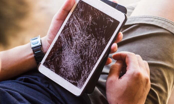 A man holds a tablet with a shattered touchscreen in one hand. He wears a blue shirt and tan shorts.