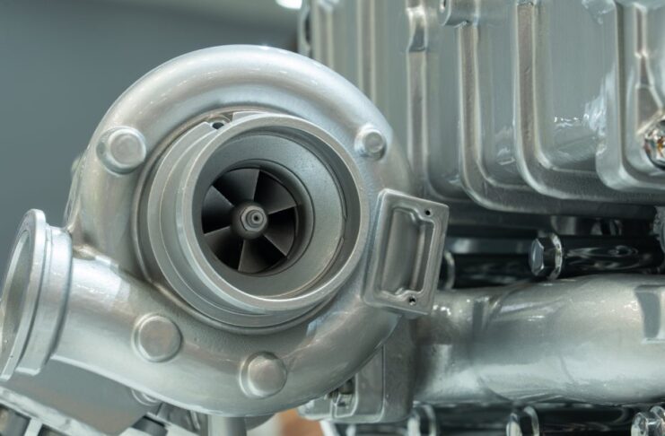 A close-up of a diesel engine turbocharger component. The part is silver, very clean, and attached to other components.