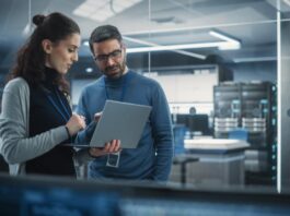 Creating a Safer Workplace in Tech Environments A man wearing eyeglasses and a woman holding a laptop stands next to each other, discussing in a server room.