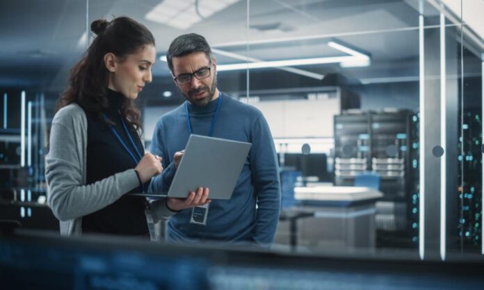 A man wearing eyeglasses and a woman holding a laptop stands next to each other, discussing in a server room.