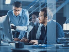 Three people wearing professional attire are looking at a large monitor in an office. One individual points at the screen.
