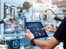 A factory worker holding a tablet in front of two white robotic arms on a brightly lit manufacturing floor.