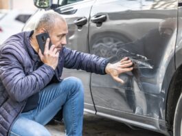 DIY Car Dent Repair: What Works and What Doesn’t A man kneeling next to the back door of his car, looking at a large scratch. He is running his hand along the scratch.
