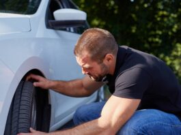 A man wearing jeans and a black T-shirt kneels next to the front tire of his white car. Both of his hands touch the tire.