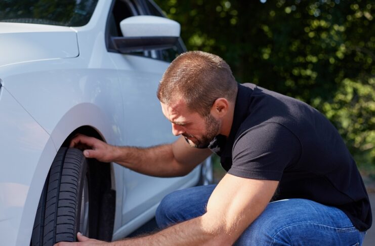 A man wearing jeans and a black T-shirt kneels next to the front tire of his white car. Both of his hands touch the tire.