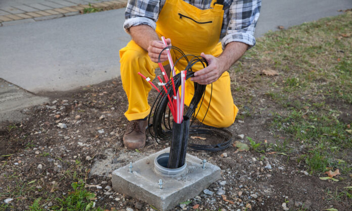 A fiber engineer wears a yellow overalls set as they kneel down to the fiber connection box on the ground.