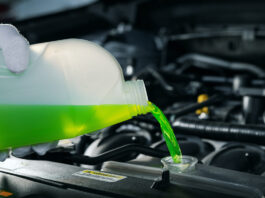 A person wearing a white glove pours bright green coolant from a translucent bottle into a car radiator.
