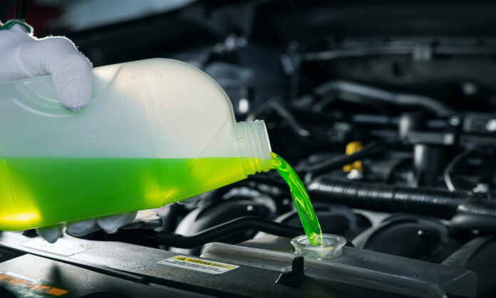 A person wearing a white glove pours bright green coolant from a translucent bottle into a car radiator.