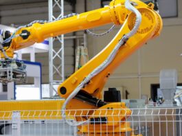 A yellow industrial robotic arm with cables and a gripper stands in a factory, surrounded by machinery and a safety fence.