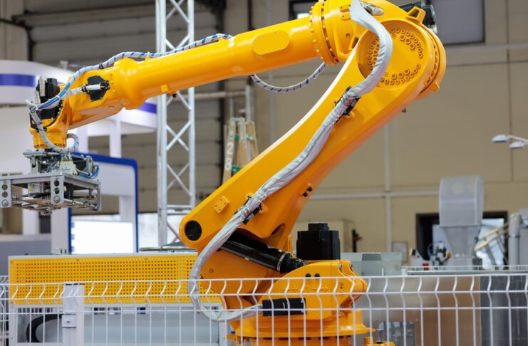 A yellow industrial robotic arm with cables and a gripper stands in a factory, surrounded by machinery and a safety fence.