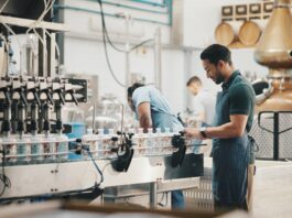 A man inspects bottles on a conveyor in a bright facility with a copper still, wooden barrels, and filling machines.