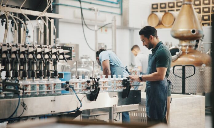 A man inspects bottles on a conveyor in a bright facility with a copper still, wooden barrels, and filling machines.