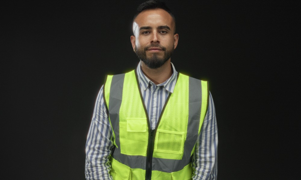 A man in a striped button-up shirt and a yellow high-visibility vest standing against a black background.