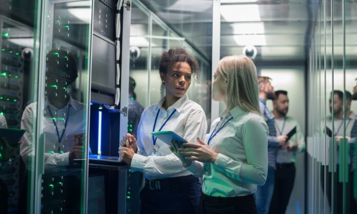 Two women stand in front of an open server rack in a data center. Others stand behind them in front of another server rack. Two women stand in front of an open server rack in a data center. Others stand behind them in front of another server rack.
