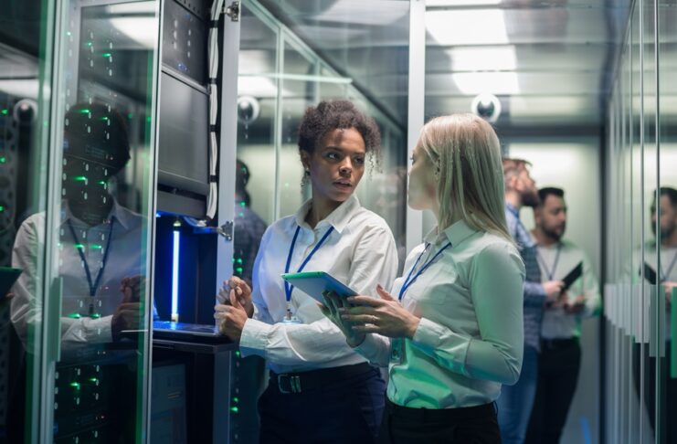 Two women stand in front of an open server rack in a data center. Others stand behind them in front of another server rack.