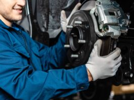 A mechanic is wearing blue coveralls and gray gloves. He's adjusting disc brakes on an elevated vehicle.