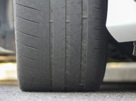 A close-up of the tire of a white car that is parked on asphalt. The tire shows uneven tread wear and faint grooves.