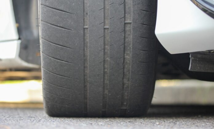 A close-up of the tire of a white car that is parked on asphalt. The tire shows uneven tread wear and faint grooves.