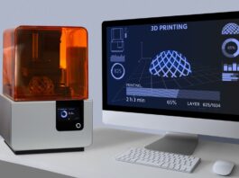 A desktop 3D printer with an orange cover sits beside a computer monitor showing a 3D print at 65% complete.