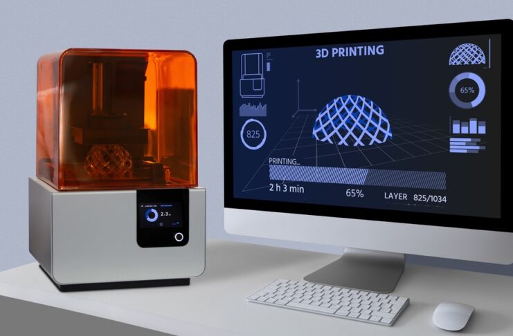 A desktop 3D printer with an orange cover sits beside a computer monitor showing a 3D print at 65% complete.