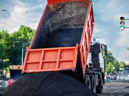 A red dump truck tilts its bed upward, releasing black asphalt onto the street at an active construction site.