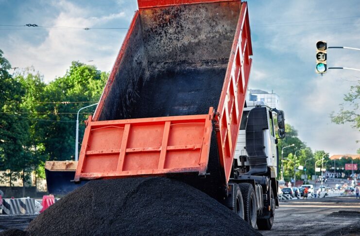 A red dump truck tilts its bed upward, releasing black asphalt onto the street at an active construction site.