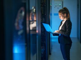 Woman in a black suit stands in a server room and types on a laptop beside server racks under blue light.