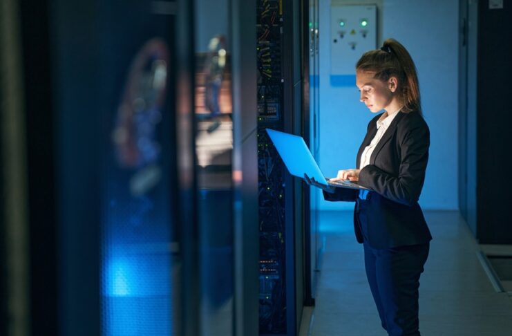 Woman in a black suit stands in a server room and types on a laptop beside server racks under blue light.