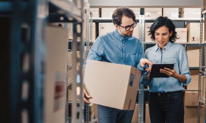 A man and woman standing in a storage room. The woman is holding a tablet and the man is holding a cardboard box.