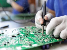 A close-up of a person wearing white gloves soldering a component onto a green printed circuit board.