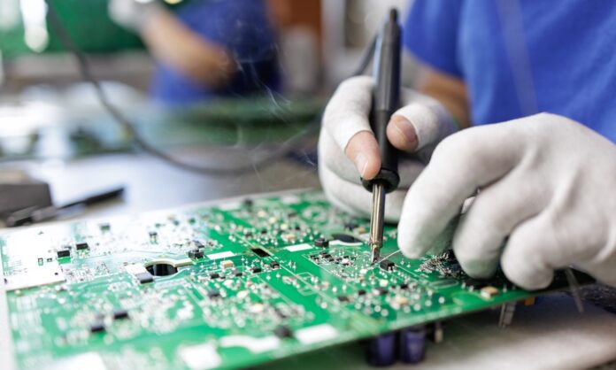 A close-up of a person wearing white gloves soldering a component onto a green printed circuit board.