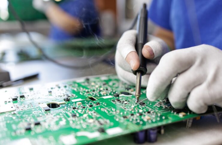 A close-up of a person wearing white gloves soldering a component onto a green printed circuit board.