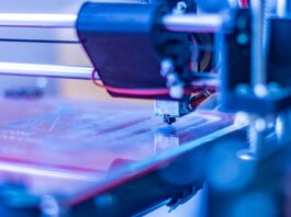 3D printer head extruding filament onto platform with mechanical arms and blue-lit workspace in background.