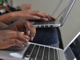 A close-up view shows two people sitting and typing on silver laptops. The laptops are sitting on their laps.