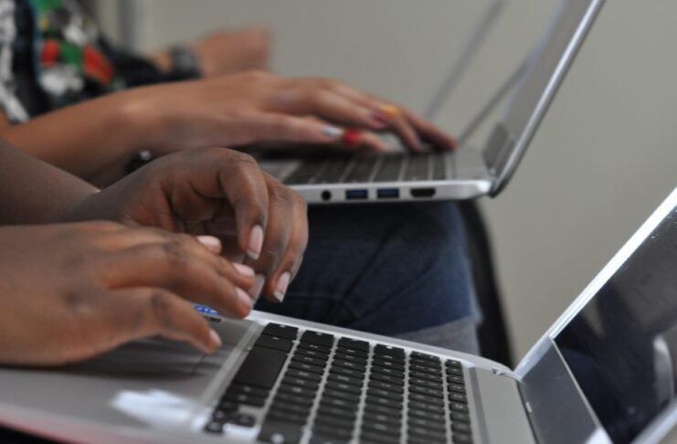 A close-up view shows two people sitting and typing on silver laptops. The laptops are sitting on their laps.