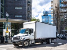 What Are Hydraulic Hybrid Vehicles, and How Do They Work? A white semi truck with a long box trailer turning at a city intersection with apartment buildings in the background.