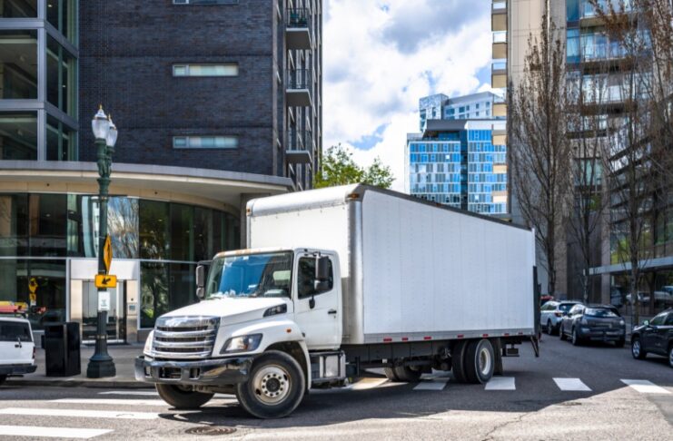 A white semi truck with a long box trailer turning at a city intersection with apartment buildings in the background.