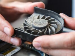 Hands hold a dust-covered laptop cooling fan, showing thick buildup on the blades inside an open computer casing.