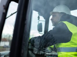 A mans its behind the wheel inside the cab of heavy machinery. He wears a yellow safety vest and a white hard hat.