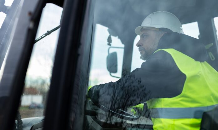 A mans its behind the wheel inside the cab of heavy machinery. He wears a yellow safety vest and a white hard hat.