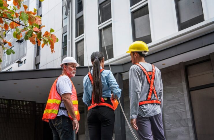 A group of engineers in safety vests and hard hats reviewing plans together at an outdoor building construction site.