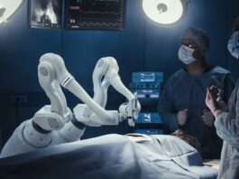 A white robotic arm holds a surgical instrument over a covered patient. Two medical staff members stand by the patient.