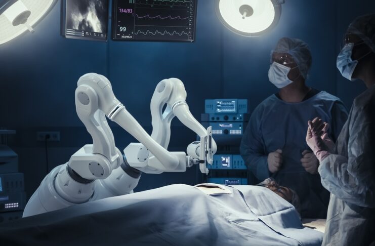 A white robotic arm holds a surgical instrument over a covered patient. Two medical staff members stand by the patient.