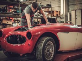 A man uses a tool to reach inside the engine bay of an older red vehicle that is being restored in a garage.