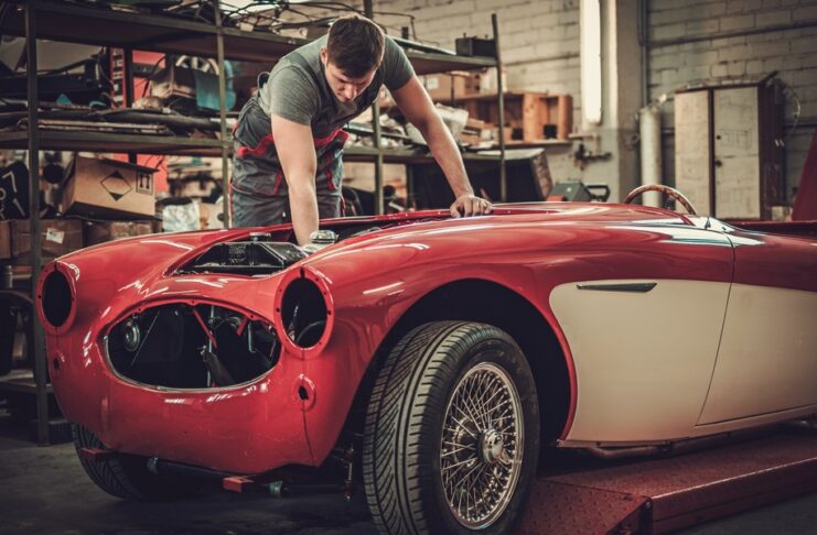 A man uses a tool to reach inside the engine bay of an older red vehicle that is being restored in a garage.