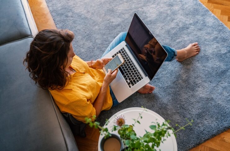 An aerial view of a female homeowner sitting on the floor in her living room with a laptop and her phone.