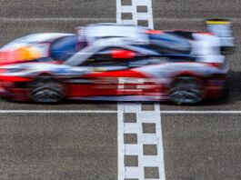 A red and white race car speeding across a finish line with motion blur, captured from above on a racetrack.